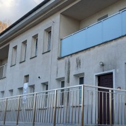 Metalsystem Barbara Brzezińska - Budynek z balkonami i złotymi barierkami. Na balkonie szkło mrożone. Widoczne okna i drzwi wejściowe. Ujęcie z dołu, niebo z chmurami w tle.