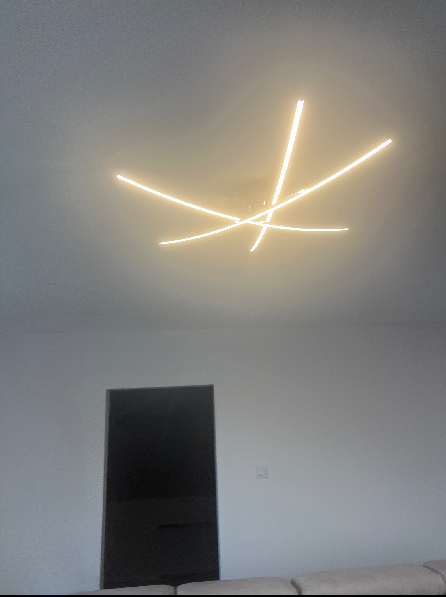 Nowoczesna lampa sufitowa LED o geometrycznym kształcie, z ciepłym światłem, w minimalistycznym wnętrzu z białymi ścianami i ciemnymi drzwiami.