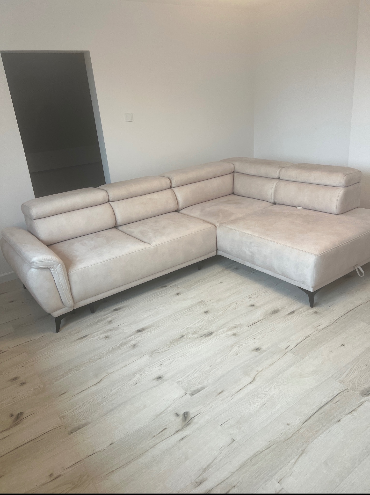 Jasny, narożny, tapicerowany zestaw wypoczynkowy z poduchami na siedziskach, ustawiony na panelach podłogowych w nowym, minimalistycznym wnętrzu.