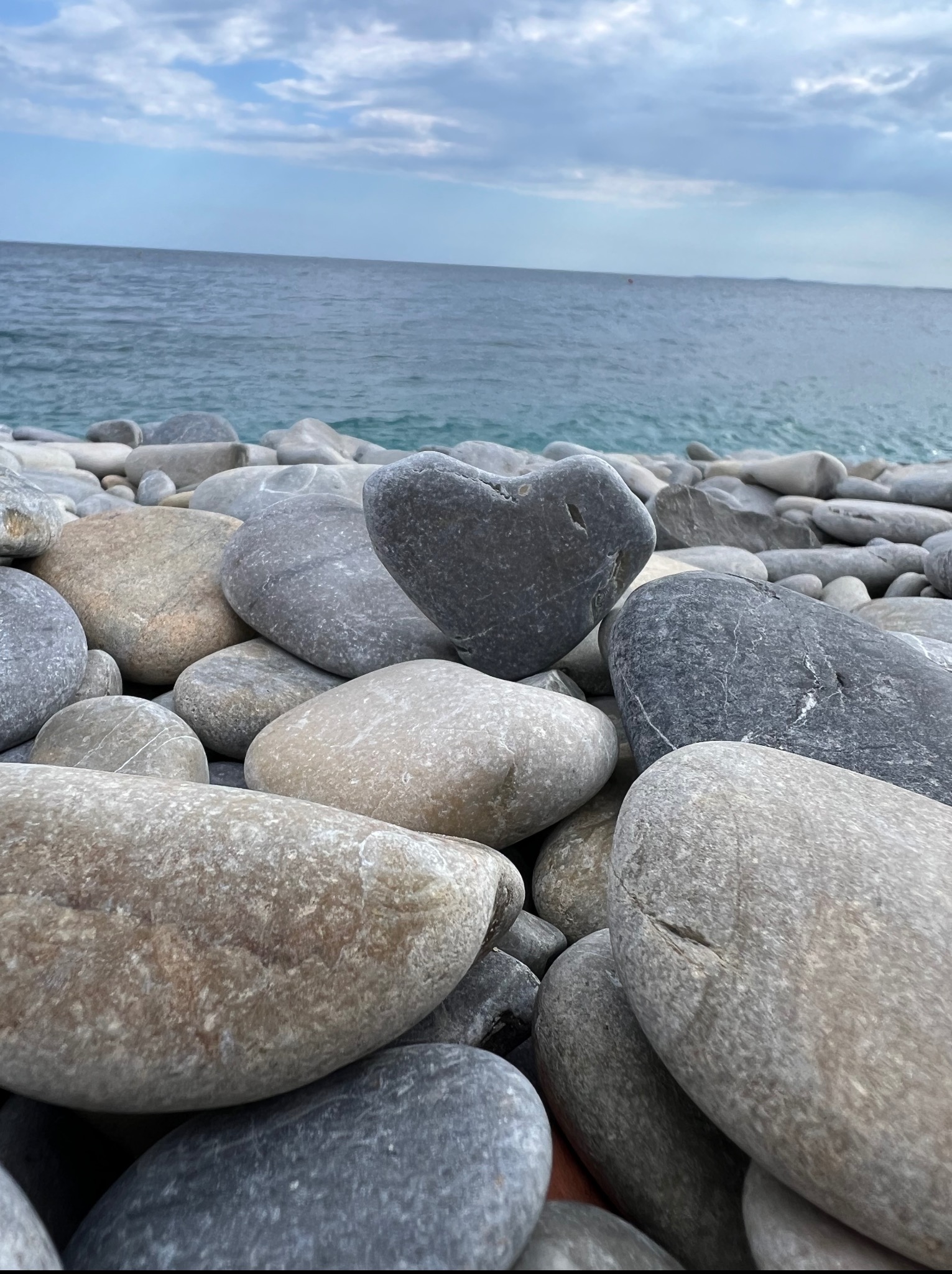 Plaża w Pułtusku z szarymi kamieniami, jeden w kształcie serca. W tle morze i pochmurne niebo. Kamienie w różnych odcieniach szarości, gładkie i okrągłe.