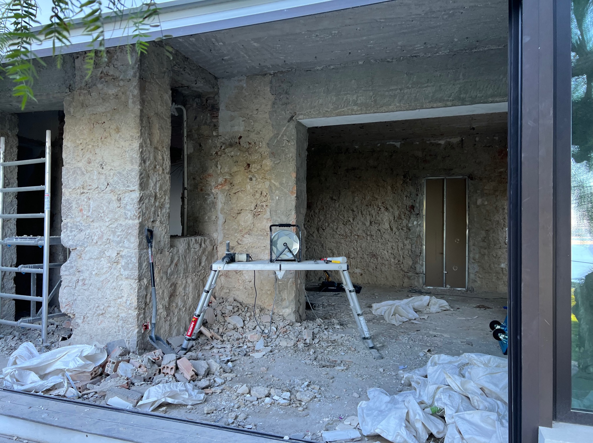 Wnętrze w trakcie remontu: surowe ściany z kamienia, gruz na podłodze, drabina, lampa na podeście. Widoczne ślady prac rozbiórkowych i przygotowania do dalszych etapów.