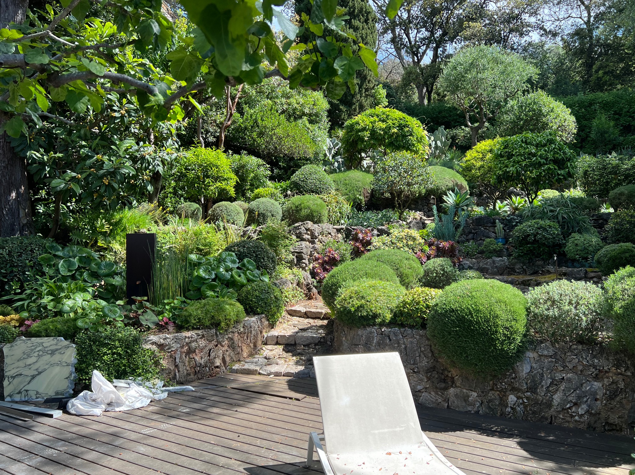 Ogród z kamiennymi ścieżkami i bujną roślinnością, widok z drewnianego tarasu z leżakiem. Marmurowe płyty i folia na tarasie sugerują prace wykończeniowe.