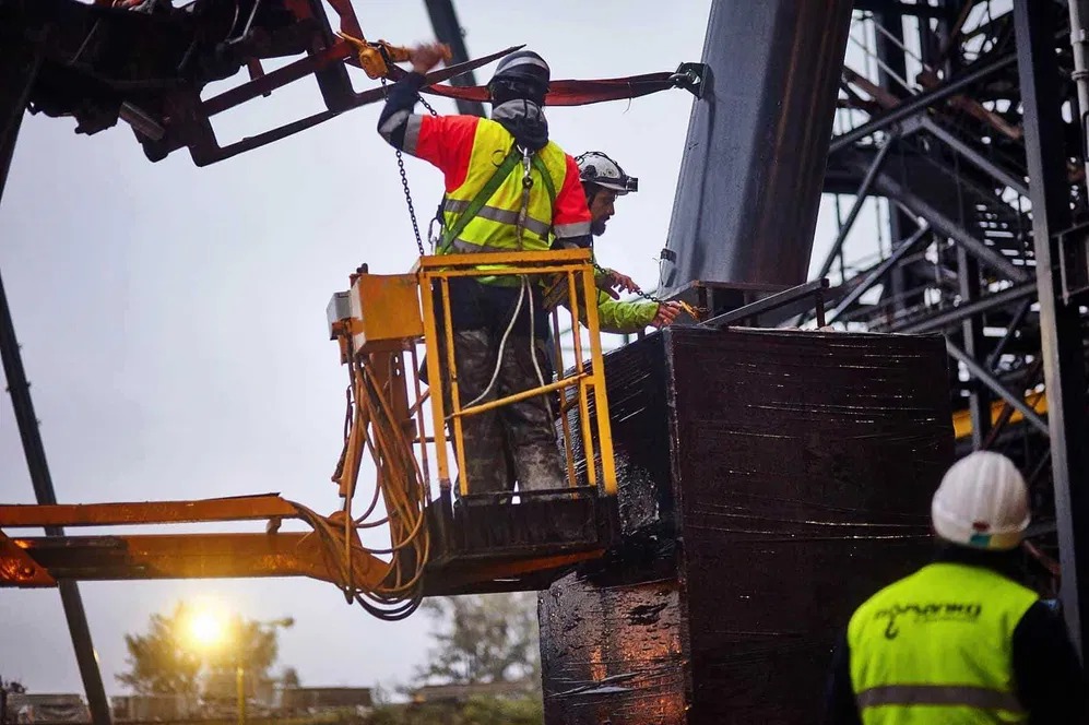 Pracownicy w koszu podnośnika koszowego, w kaskach i odblaskowych ubraniach, montują element konstrukcyjny na wysokości. Widoczny fragment żurawia i konstrukcji stalowej.