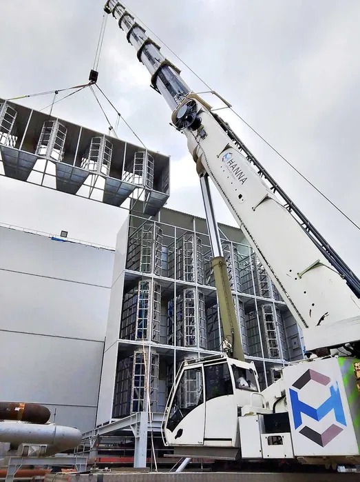 Żuraw samojezdny podnosi modułową konstrukcję na tle budynku. Widoczny fragment żurawia z logo firmy Hanna. Pochmurne niebo w tle.