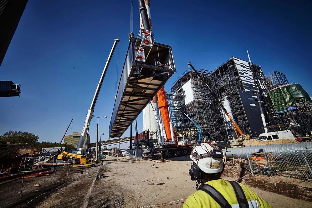 Montaż konstrukcji stalowej żurawiem na budowie w Warszawie. Widoczny pracownik w kasku i kamizelce odblaskowej, budynek w rusztowaniach i błękitne niebo.