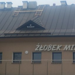 MARBUD - Ciemny dach budynku z drewnianą konstrukcją na górze, prawdopodobnie w trakcie naprawy lub wymiany. Widoczny fragment budynku z napisem 'ŻŁOBEK MIE'
