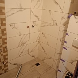 MARBUD - Układanie płytek w łazience: białe płytki imitujące marmur na ścianach i podłodze. Widoczne poziomica, otwory na armaturę i fragment instalacji. Prace w toku.