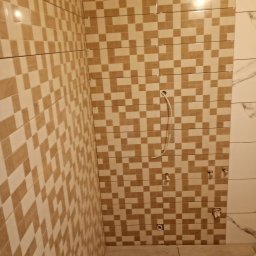 MARBUD - Ukończona ściana łazienki z mozaiką w odcieniach beżu i brązu, widoczne przyłącza wodne i fragment podłogi z odpływem liniowym. Część ściany wykończona płytami imitującymi marmur.