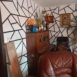 MARBUD - Pokój z geometrycznym wzorem na ścianie, drewnianym pianinem i brązowym fotelem. Widoczne panele podłogowe, rośliny doniczkowe i dekoracje.