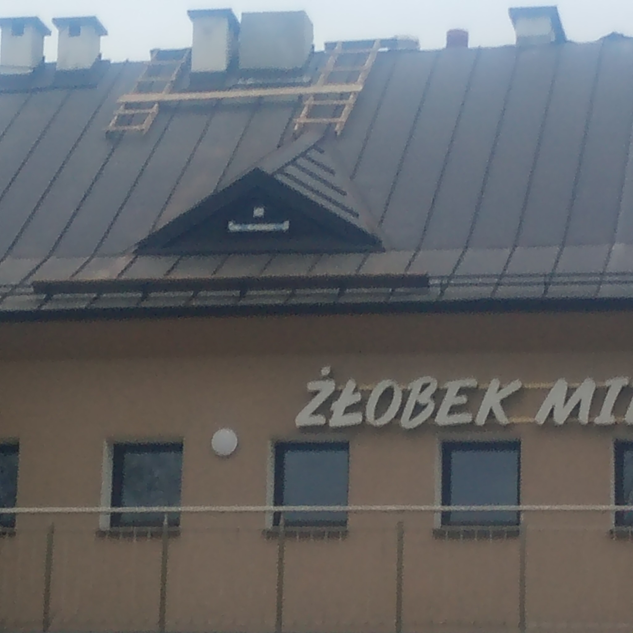 Ciemny dach budynku z drewnianą konstrukcją na górze, prawdopodobnie w trakcie naprawy lub wymiany. Widoczny fragment budynku z napisem 'ŻŁOBEK MIE'