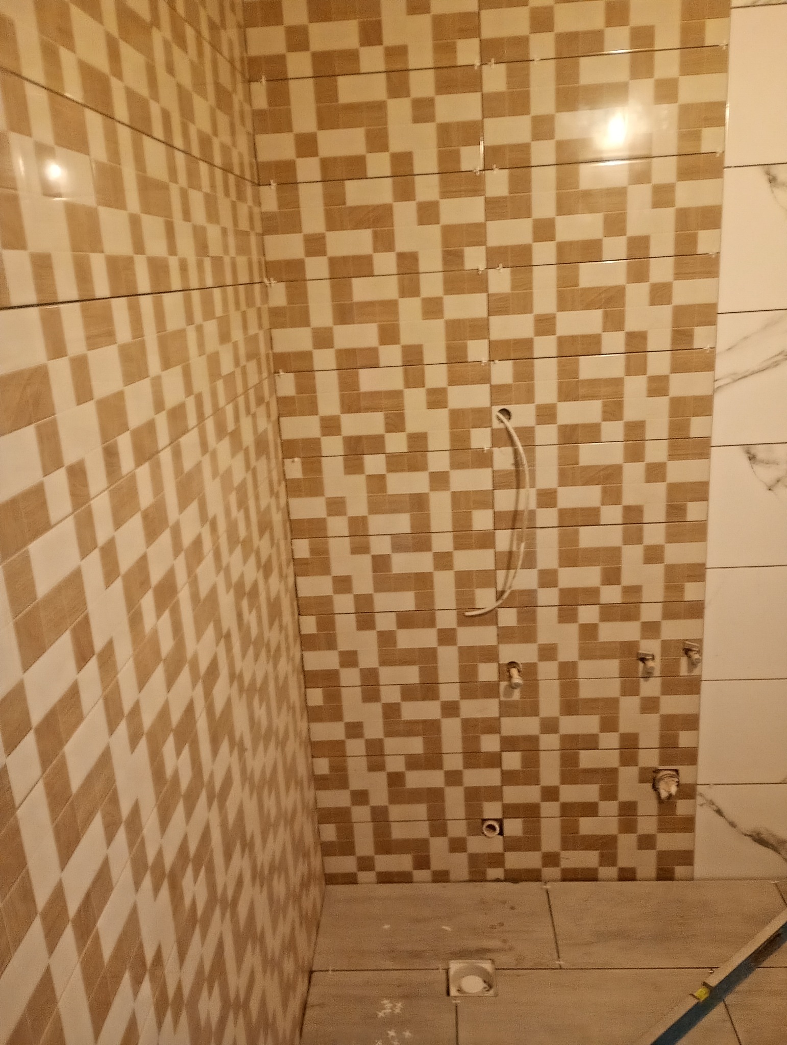 Ukończona ściana łazienki z mozaiką w odcieniach beżu i brązu, widoczne przyłącza wodne i fragment podłogi z odpływem liniowym. Część ściany wykończona płytami imitującymi marmur.