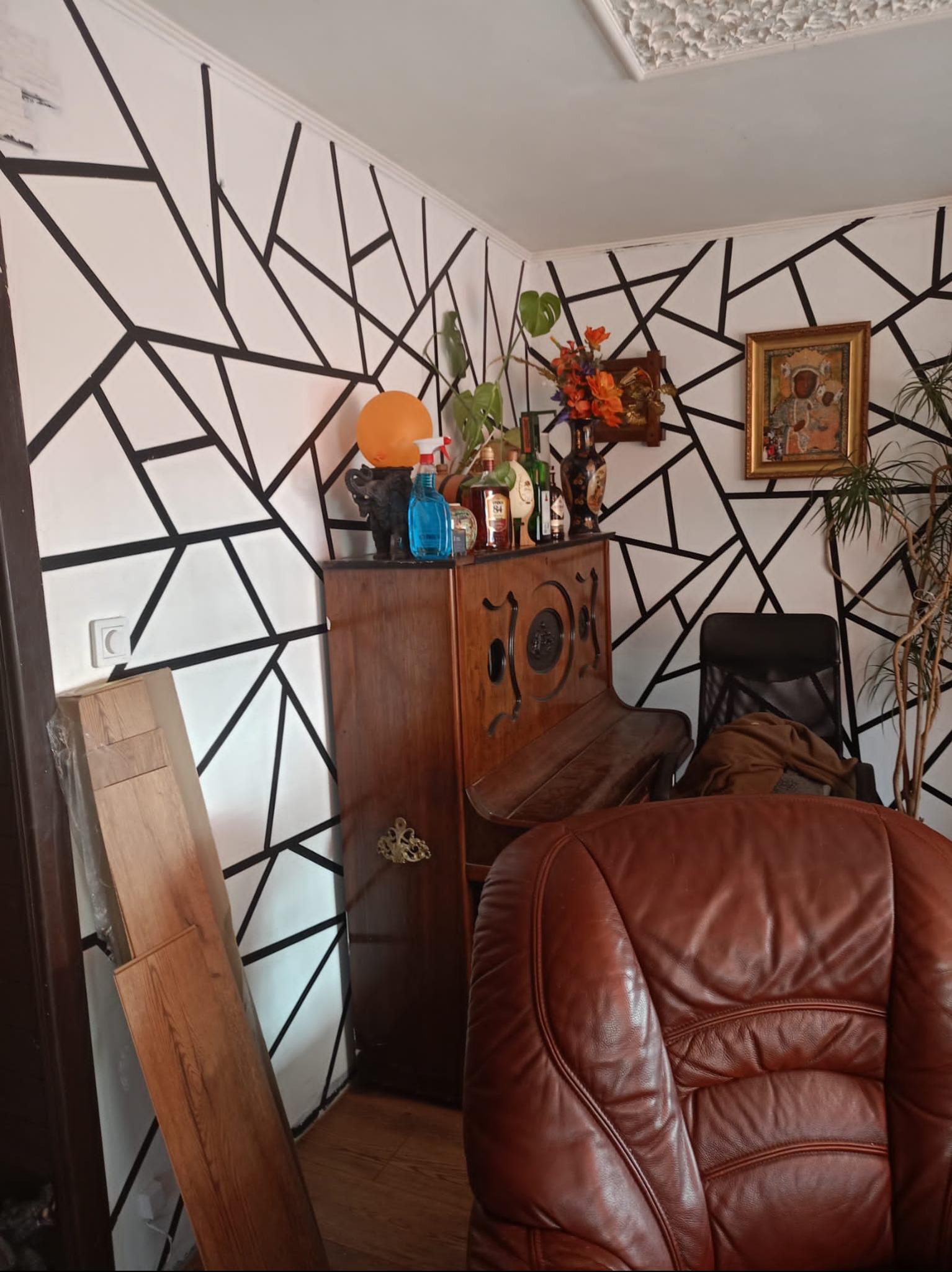 Pokój z geometrycznym wzorem na ścianie, drewnianym pianinem i brązowym fotelem. Widoczne panele podłogowe, rośliny doniczkowe i dekoracje.