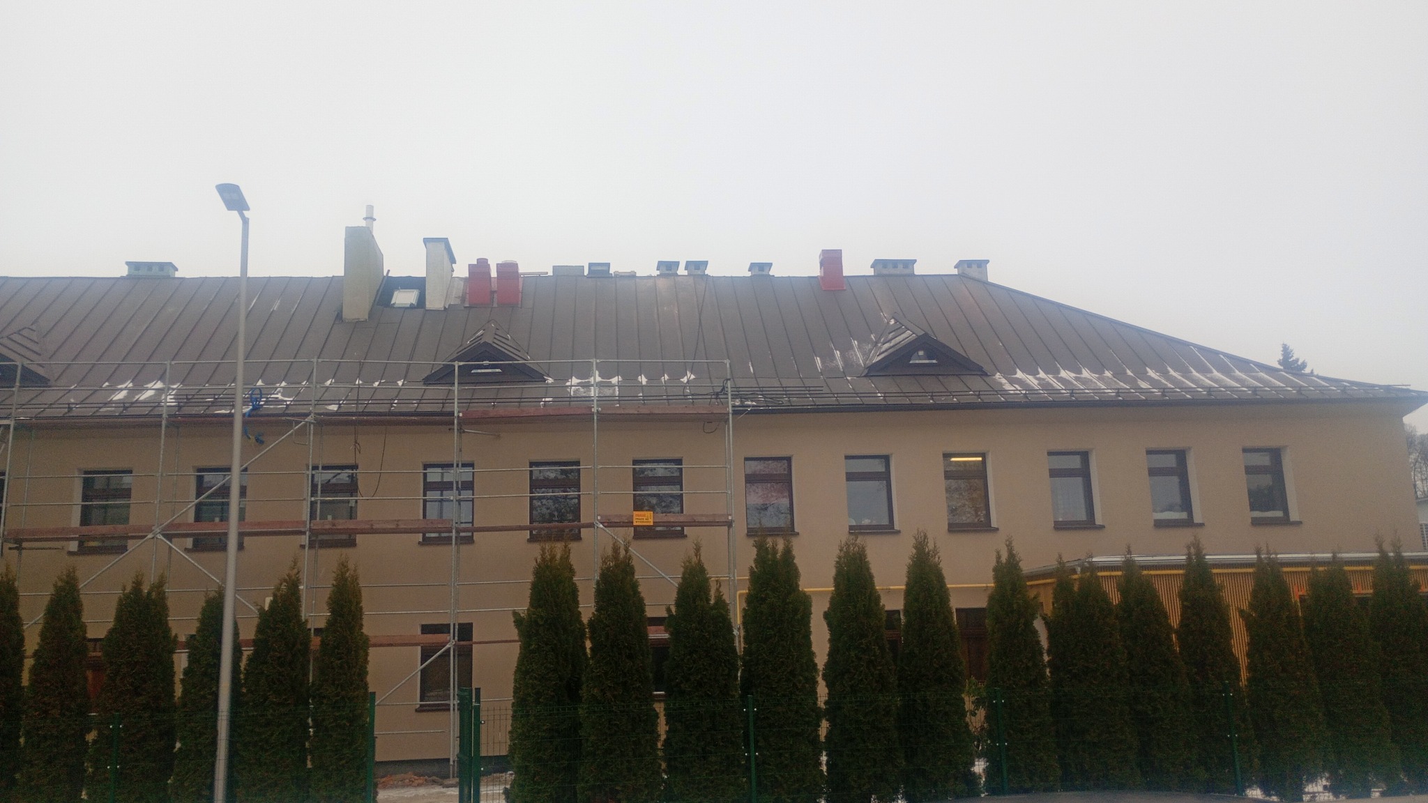 Budynek z szarym dachem, częściowo pokryty rusztowaniem. Widoczne kominy, okna i rząd zielonych drzew iglastych. Pochmurne niebo w tle.