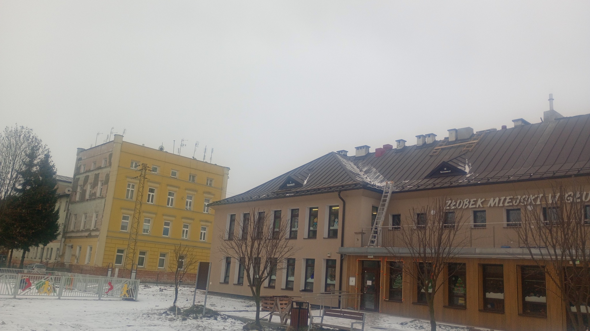 Żłobek Miejski w zimowej scenerii z częściowo zdjętym dachem, drabina oparta o budynek. Widoczne elementy konstrukcji dachu podczas naprawy.