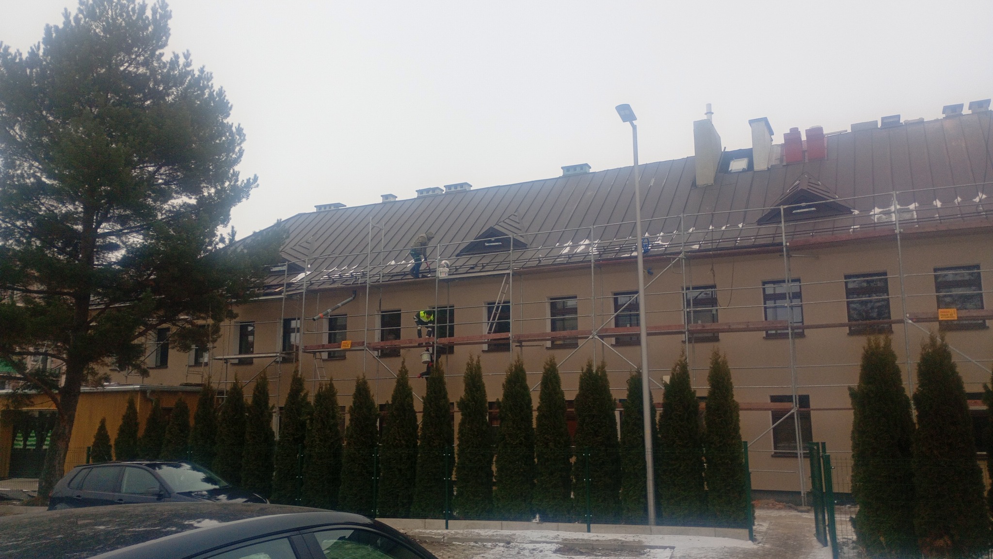 Praca na dachu budynku z blachy trapezowej, widoczni dekarze na rusztowaniu. Częściowo ośnieżony dach i otoczenie. W tle drzewa iglaste i budynek z beżową elewacją.
