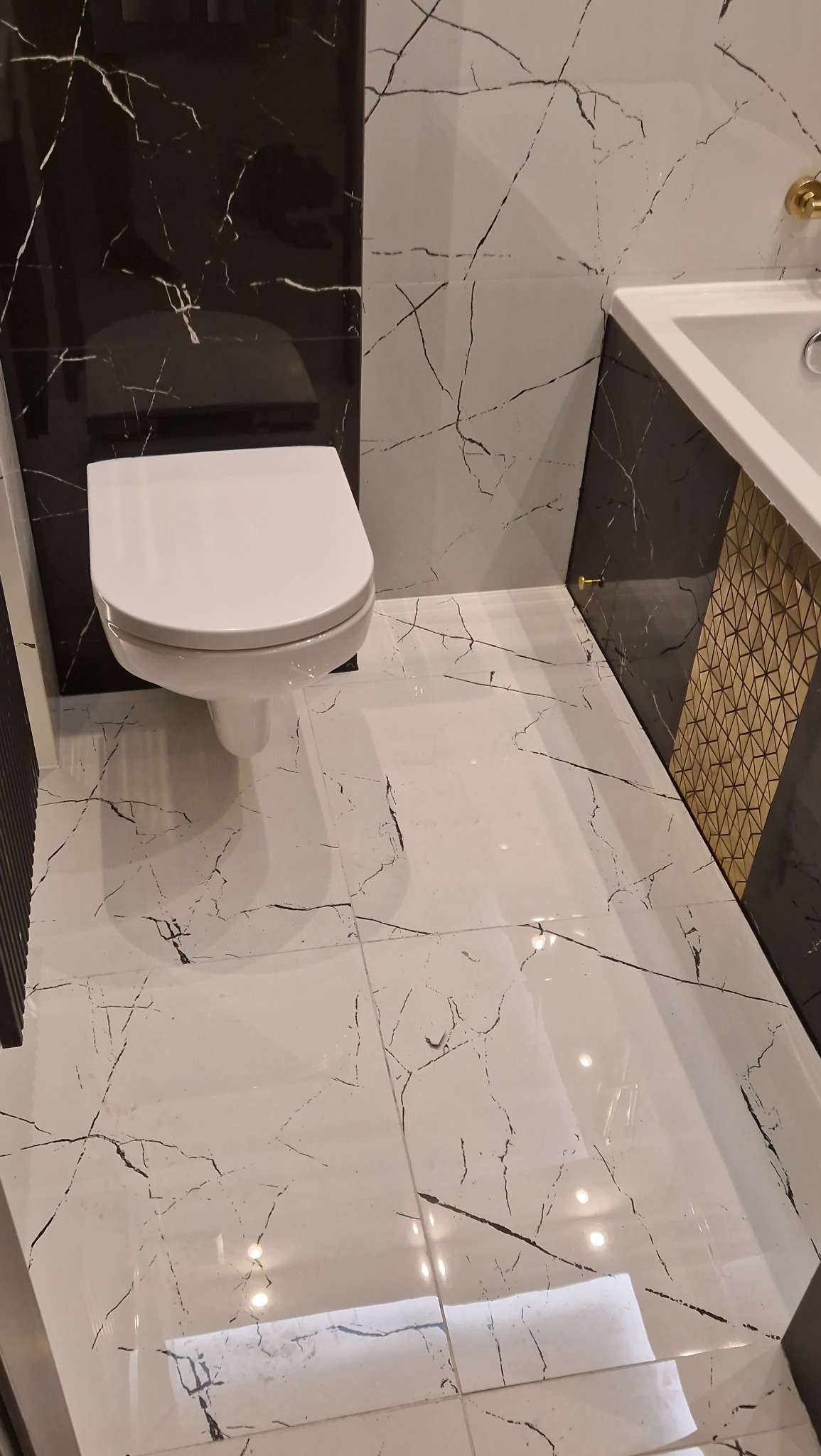 Nowoczesna toaleta w Toruniu z białymi płytkami imitującymi marmur z czarnymi żyłami, kontrastującymi z czarną ścianą i złotymi akcentami przy umywalce.