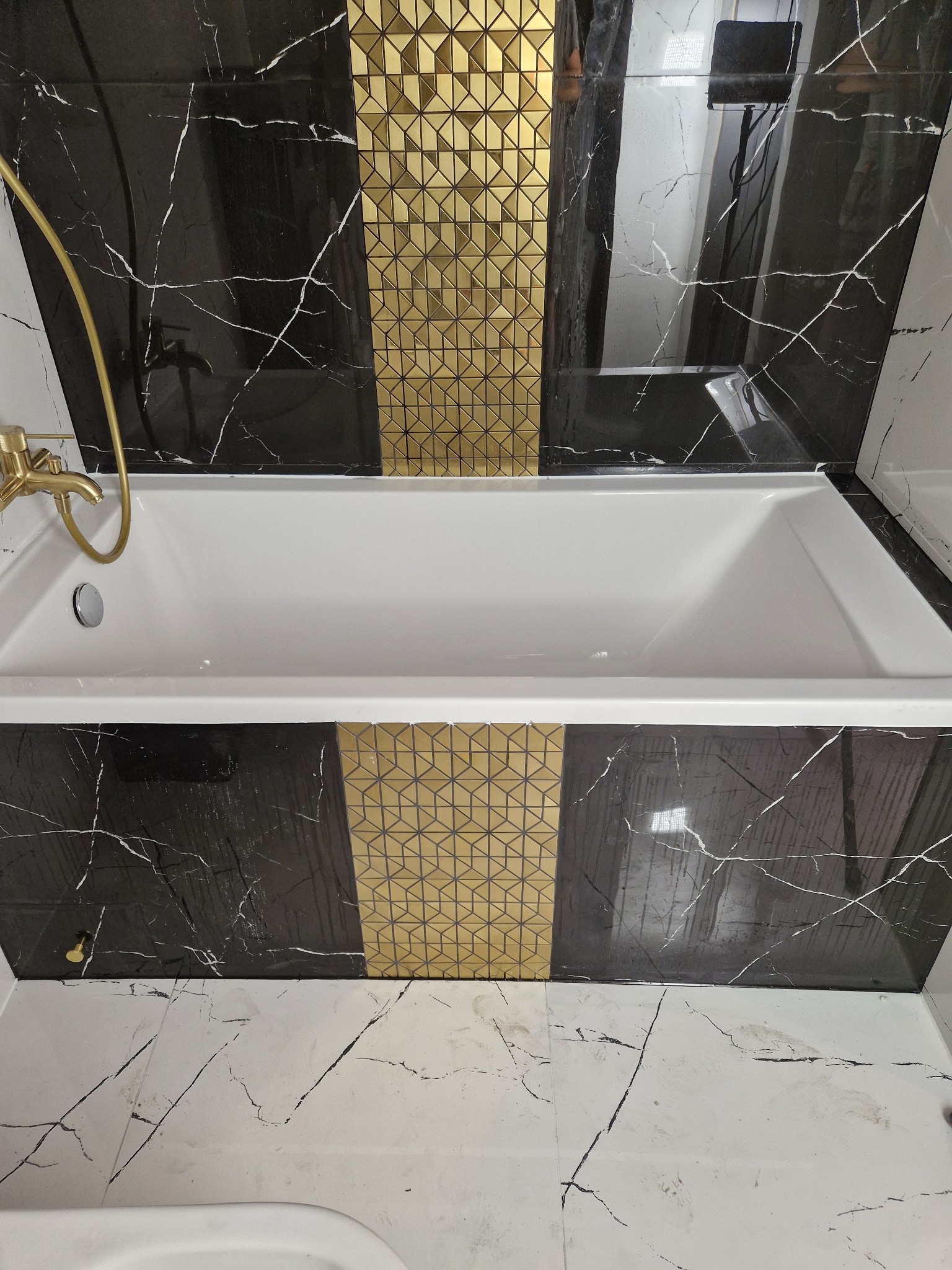 Biała wanna z baterią, otoczona czarnymi płytkami imitującymi marmur i złotymi mozaikami w łazience. Wykończenie z dbałością o detale.