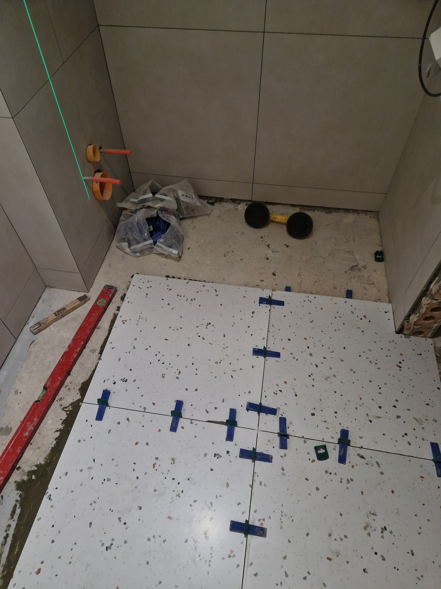 Układanie białych płytek podłogowych z kolorowym wzorem, z użyciem niebieskich krzyżyków dystansowych i poziomicy. Widoczne rury instalacyjne w ścianie.