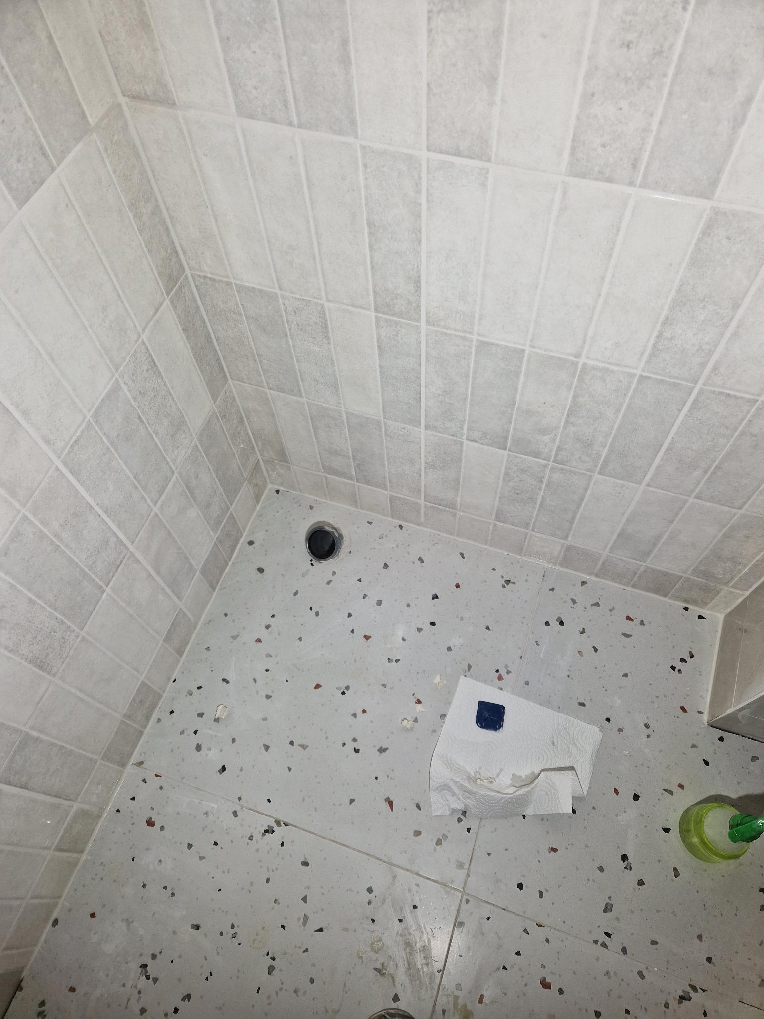 Wykończona kabina prysznicowa z jasnymi, kwadratowymi płytkami na ścianach i podłogą w stylu lastryko. Widoczny odpływ i akcesoria czyszczące.