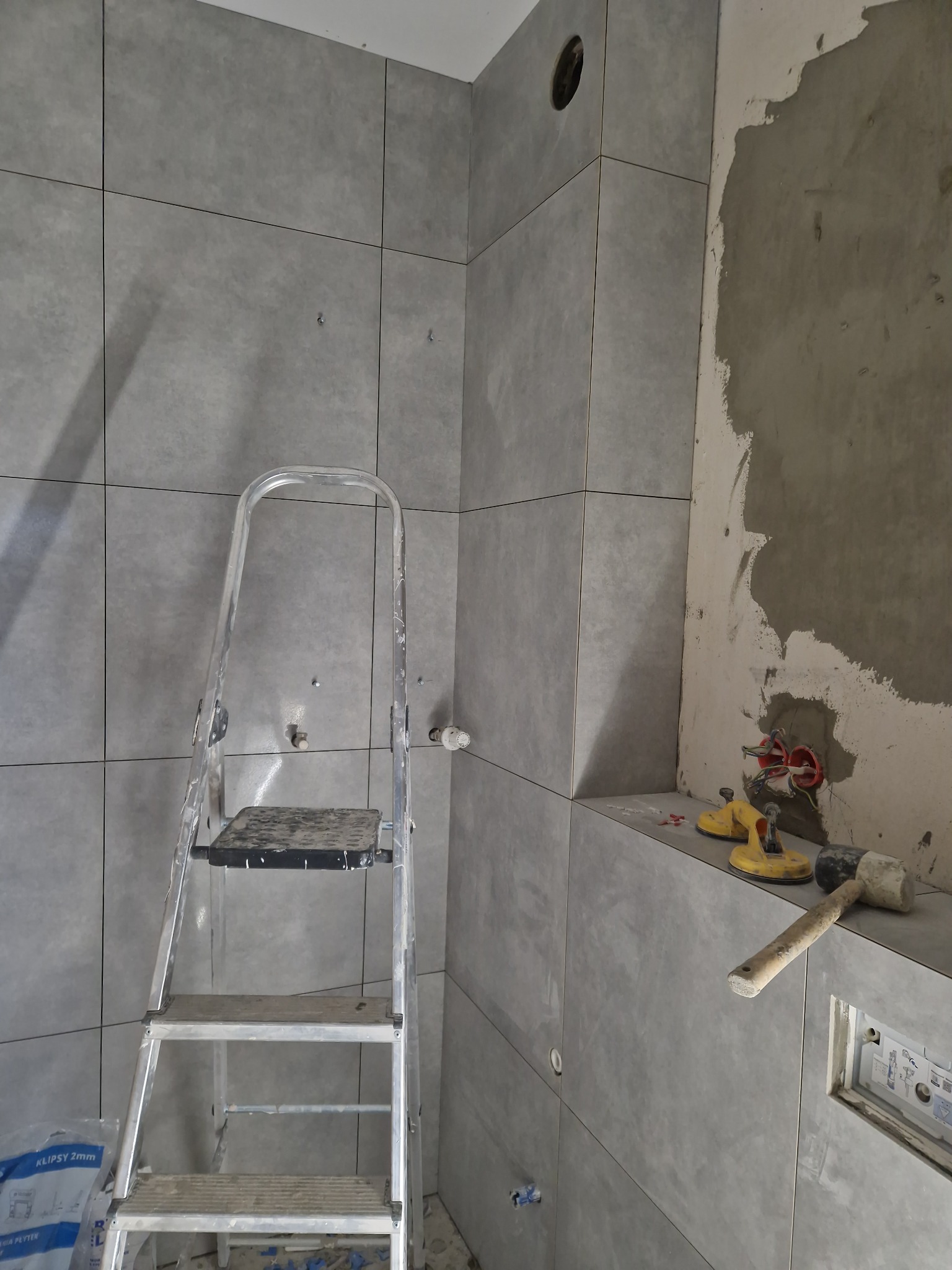 Szare płytki ścienne w łazience w trakcie montażu, widoczna drabina, przyssawki do płytek i młotek gumowy. Częściowo wykończona ściana z instalacją elektryczną.