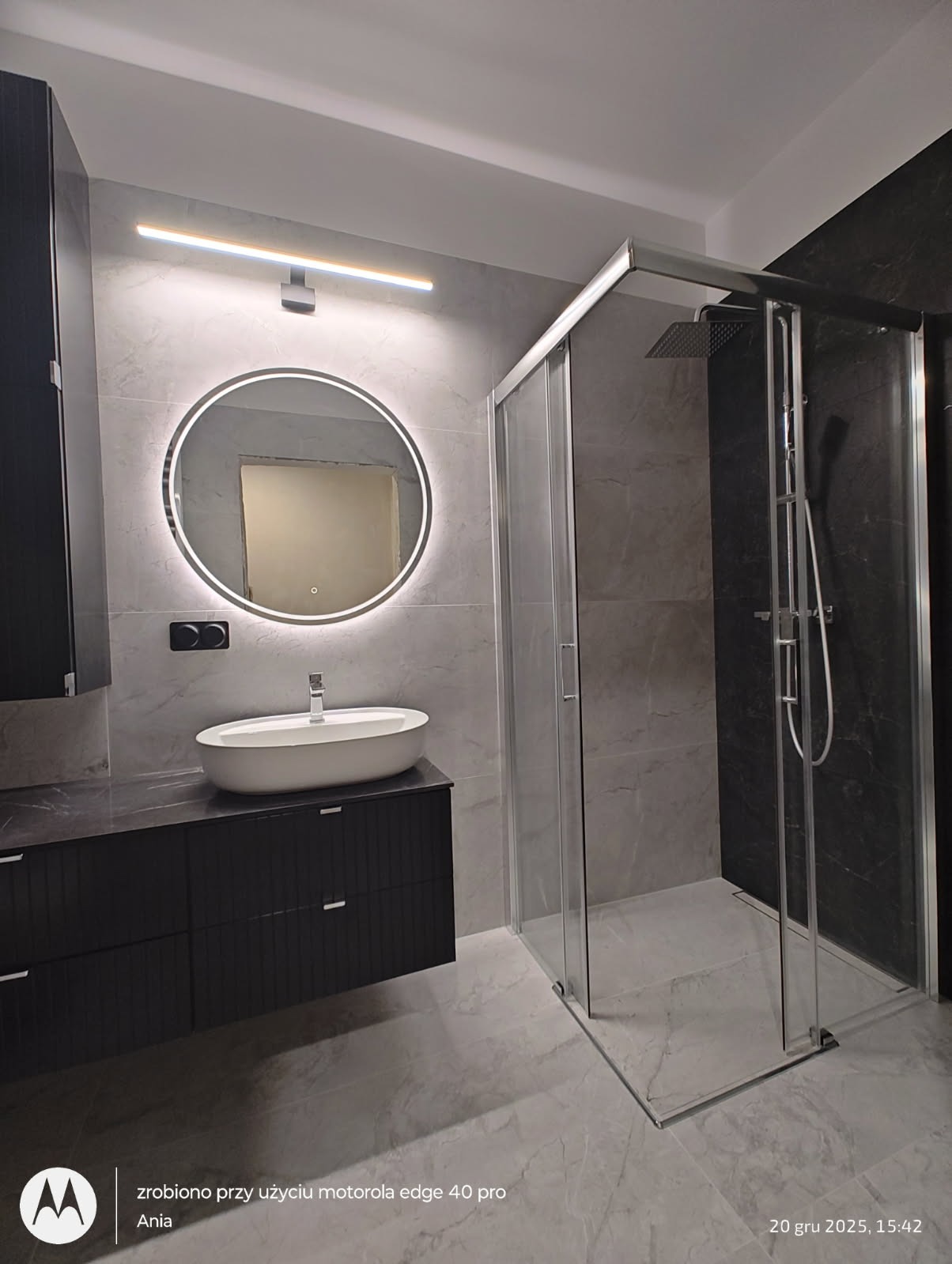 Nowoczesna łazienka z owalną umywalką, podświetlanym lustrem i kabiną prysznicową typu walk-in. Szare płytki imitujące marmur na ścianach i podłodze.