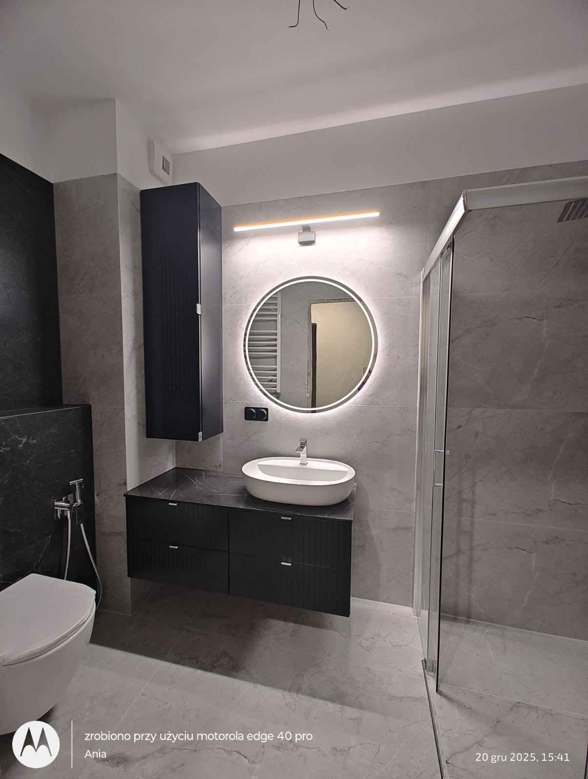 Nowoczesna łazienka w szarościach z okrągłym lustrem LED, czarną szafką pod umywalką i kabiną prysznicową. Minimalistyczny design, marmurowe płytki i oświetlenie.