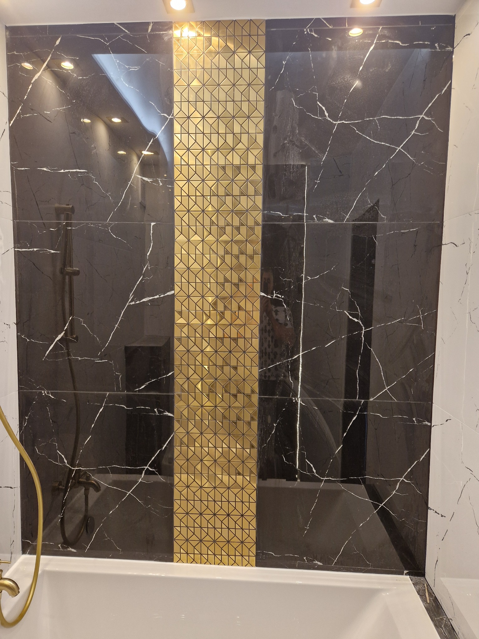 Wykończona ściana nad wanną z czarnymi płytkami imitującymi marmur, przedzielona pionowym pasem złotej mozaiki. Widoczna bateria wannowa i fragment kabiny prysznicowej.