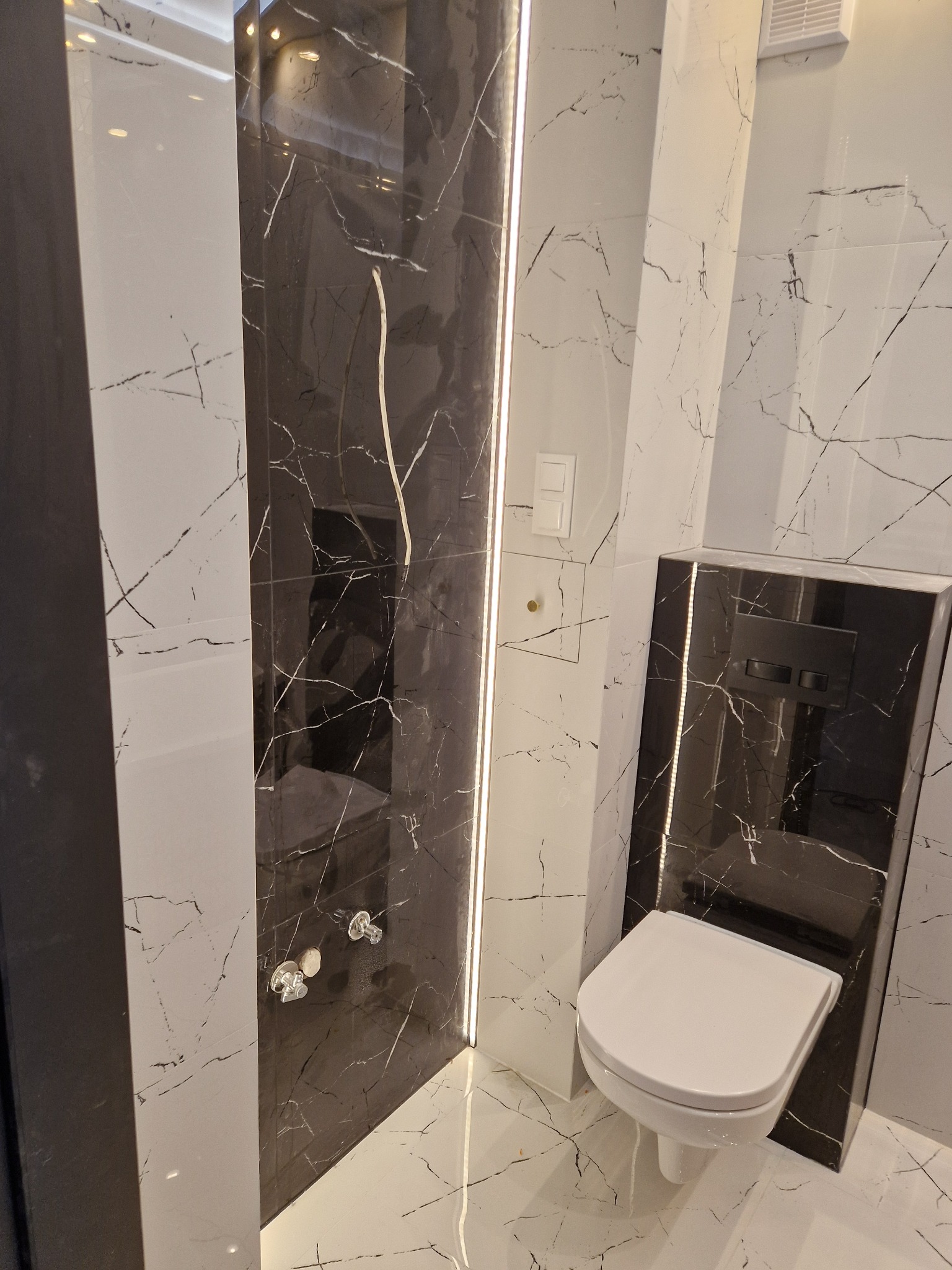 Nowoczesna łazienka z marmurowymi płytkami w odcieniach bieli i czerni, podświetleniem LED i podwieszaną toaletą. Minimalistyczny design z eleganckimi detalami.