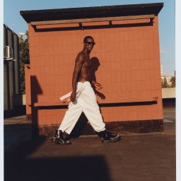 NO PLANET B SPÓŁKA Z OGRANICZONĄ ODPOWIEDZIALNOŚCIĄ - Mężczyzna w okularach przeciwsłonecznych, bez koszuli, w białych spodniach i czarnych butach, idzie na tle ceglanej ściany w słoneczny dzień. Widoczne budynki w tle.