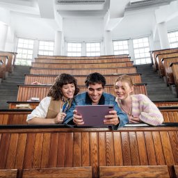 NO PLANET B SPÓŁKA Z OGRANICZONĄ ODPOWIEDZIALNOŚCIĄ - Trzy osoby w auli uniwersyteckiej oglądają tablet Huawei. Uśmiechnięci, siedzą w ławkach, z widocznymi schodami i oknami w tle. Jasne oświetlenie, ciepłe barwy drewna.