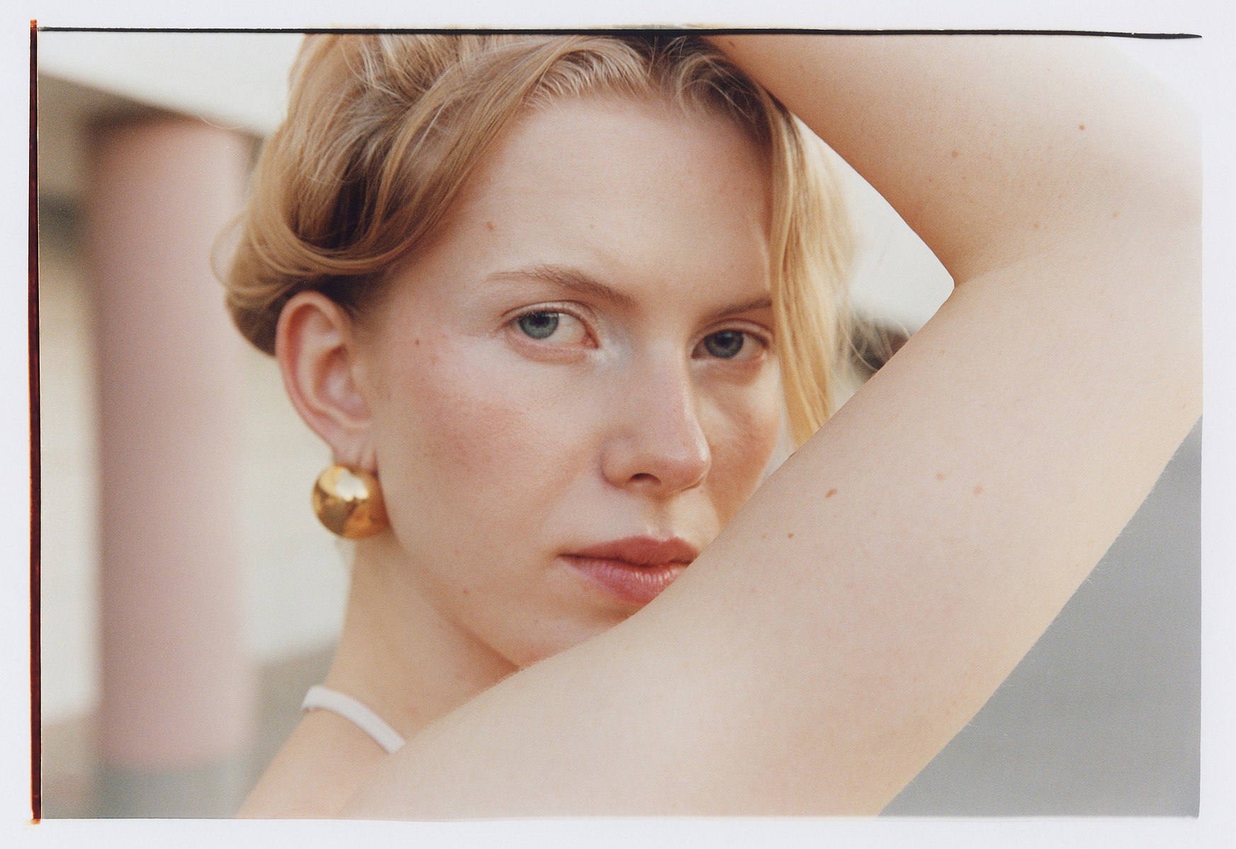 Portret kobiety z blond włosami i złotymi kolczykami, ramię uniesione nad głową, delikatny makijaż, naturalne światło, minimalistyczne tło.