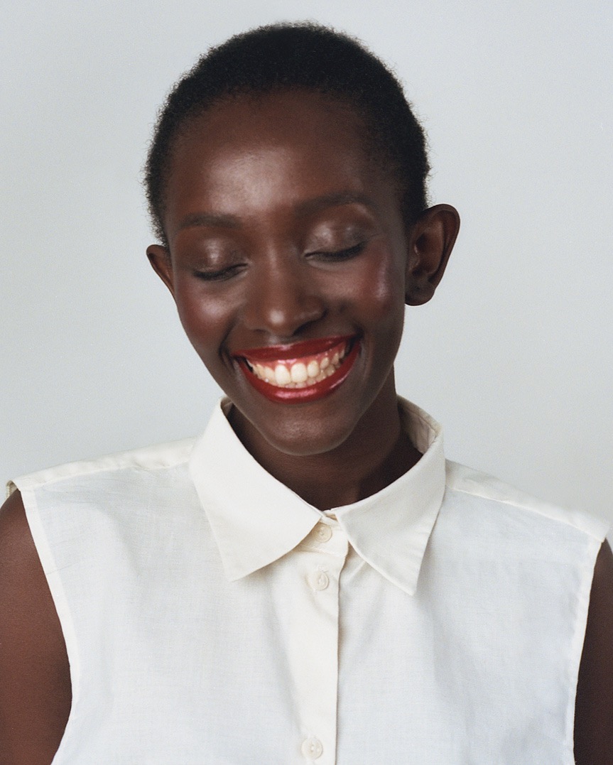 Uśmiechnięta, ciemnoskóra kobieta z zamkniętymi oczami, w białej koszuli bez rękawów, z intensywnie czerwoną szminką na ustach, na jasnym tle.