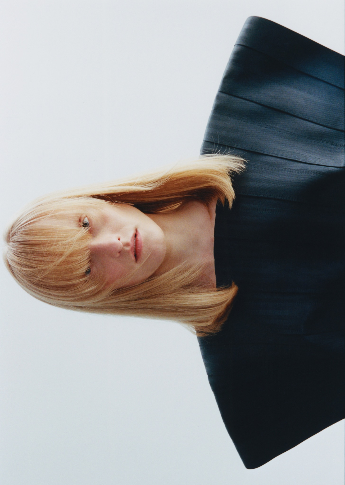 Portret kobiety z blond włosami i ciemną, geometryczną bluzką na jasnym tle, uchwycony pod nietypowym kątem, tworzący minimalistyczną kompozycję.