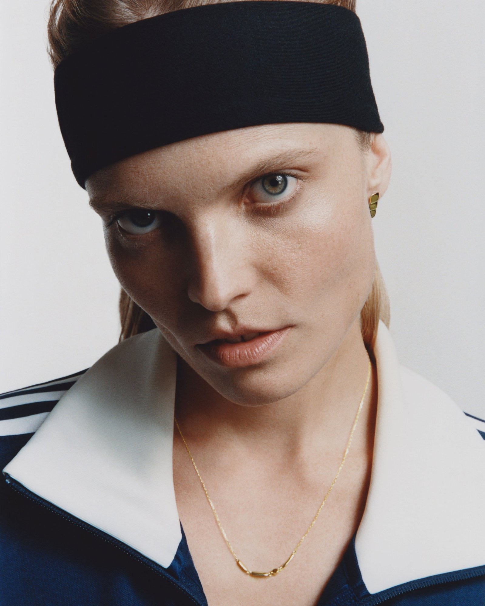 Portret kobiety z czarną opaską na włosach, w sportowej bluzie z trzema paskami, z delikatnym złotym łańcuszkiem i kolczykiem. Ujęcie z bliska, ostre światło.