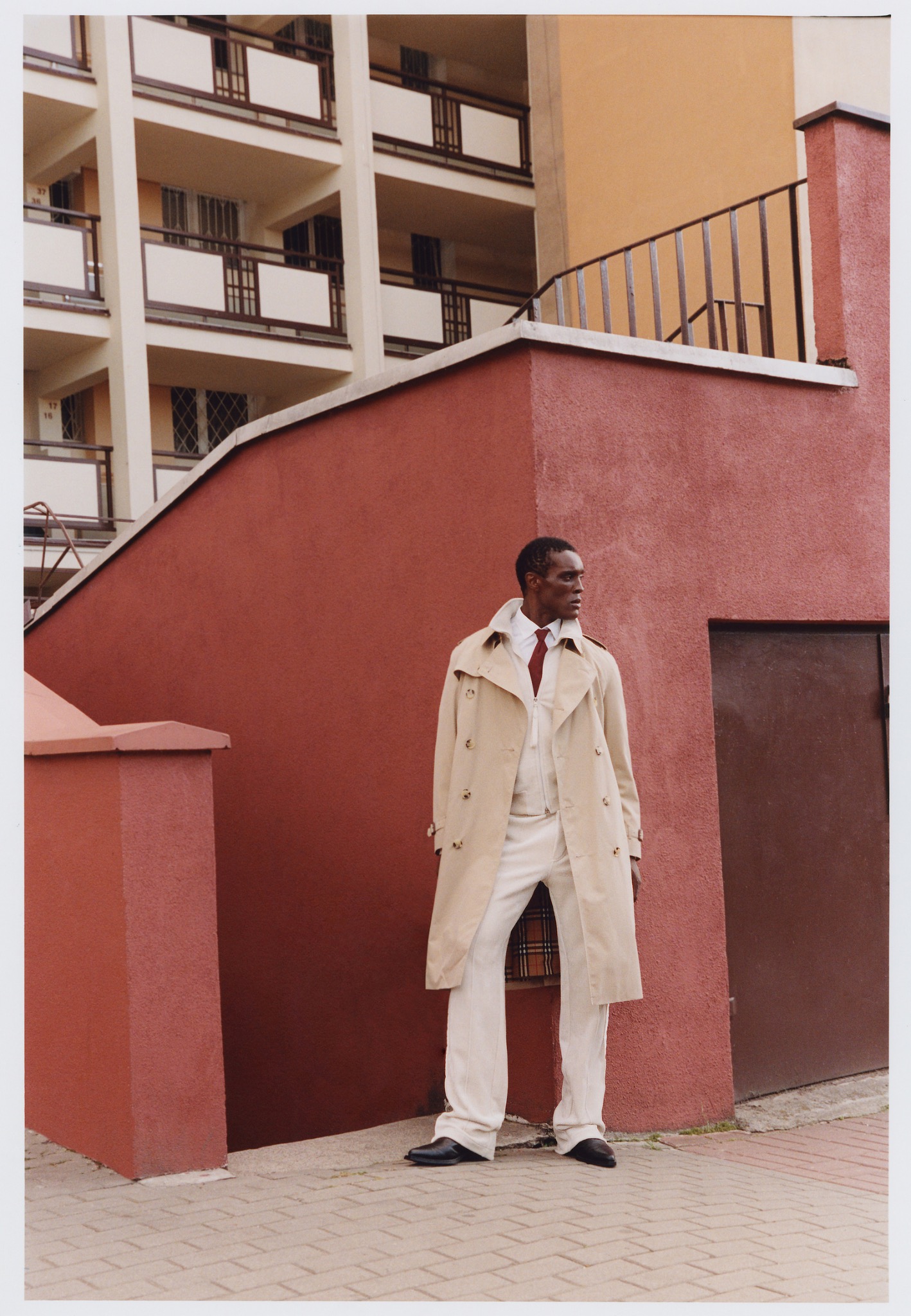 Mężczyzna w beżowym trenczu, białym garniturze i czerwonym krawacie pozuje na tle bloku z czerwonej cegły. Ujęcie z dołu, podkreślające architekturę i stylizację.