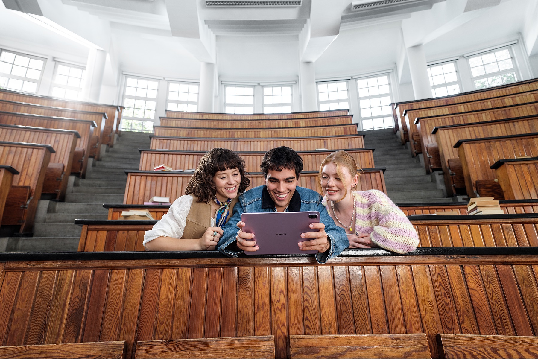 Trzy osoby w auli uniwersyteckiej oglądają tablet Huawei. Uśmiechnięci, siedzą w ławkach, z widocznymi schodami i oknami w tle. Jasne oświetlenie, ciepłe barwy drewna.