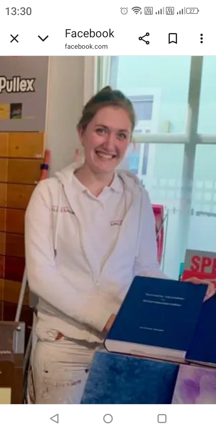 Uśmiechnięta malarka w ubraniu roboczym, prezentuje album z realizacjami na tle okna i materiałów malarskich. Widoczny logotyp firmy na bluzie.