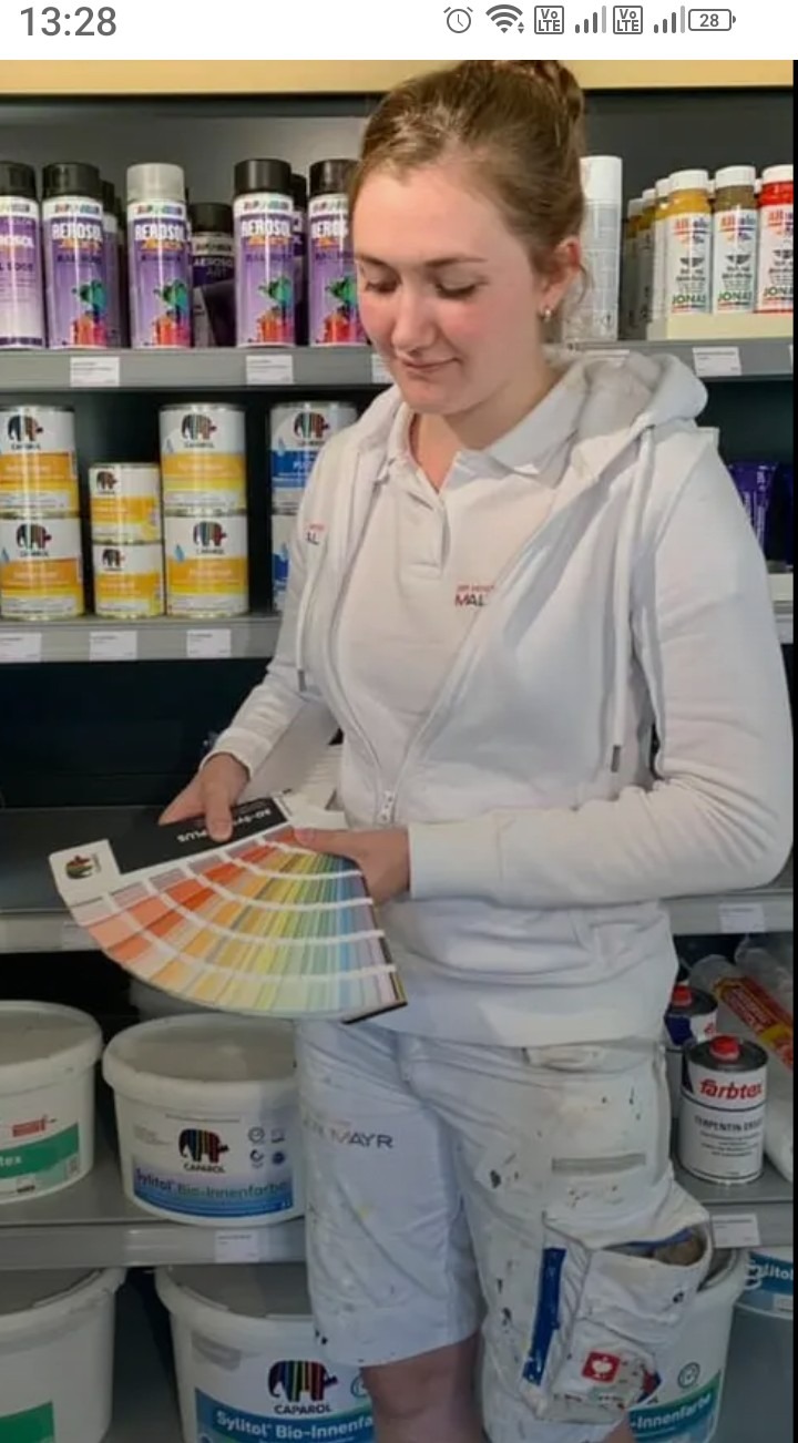 Młoda kobieta w białym stroju roboczym trzyma wachlarz z próbkami kolorów farb na tle półek z puszkami farb w sklepie z artykułami malarskimi.