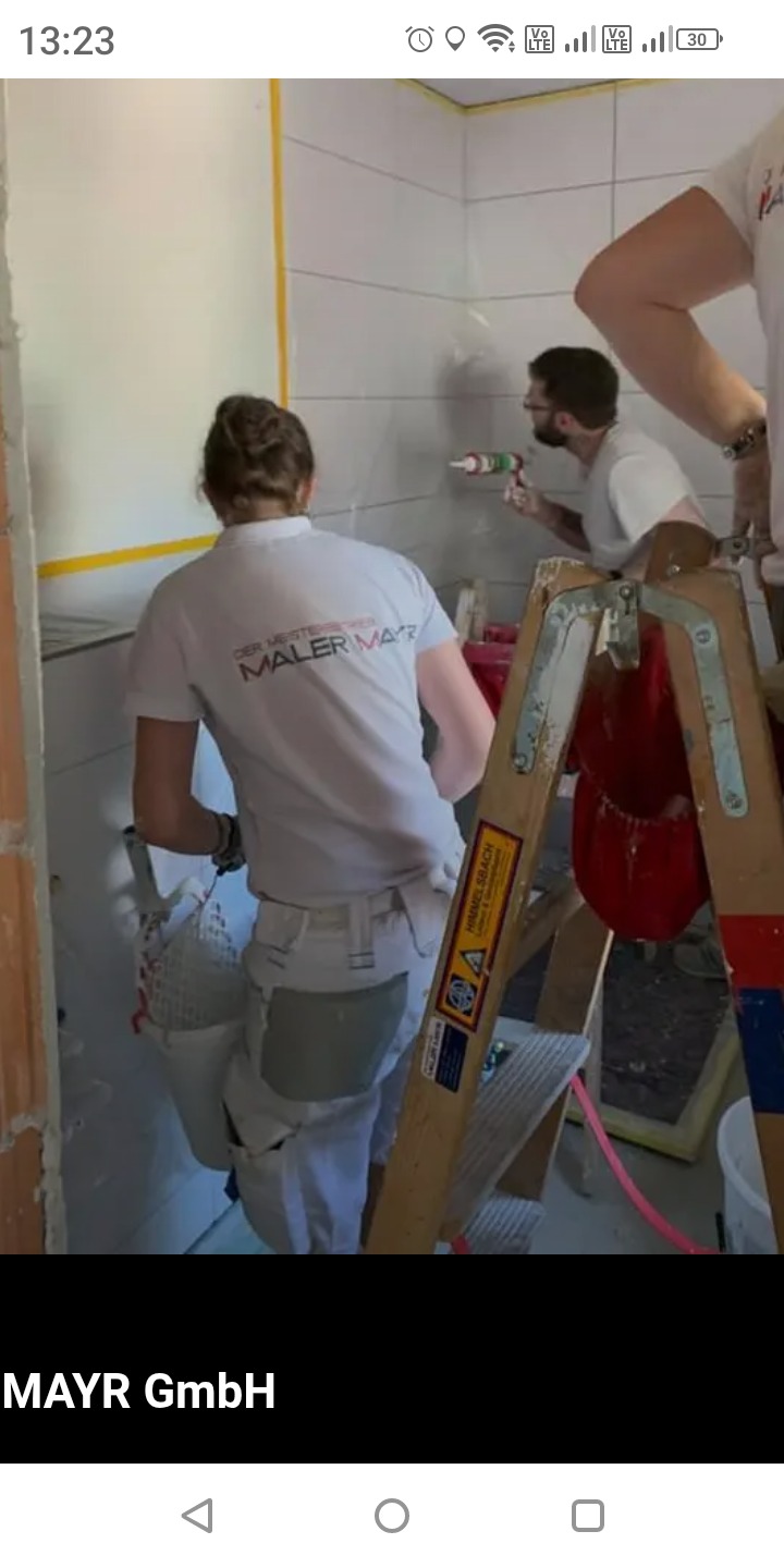 Pracownicy w trakcie spoinowania płytek w łazience. Jeden z nich używa silikonu, drugi przygotowuje się do pracy. Widoczna drabina i narzędzia.