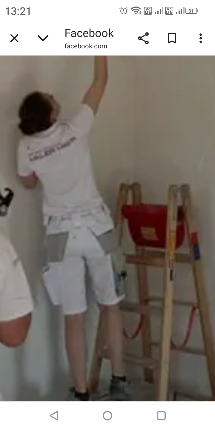 Malarz w białym stroju roboczym na drabinie maluje ścianę w rogu pomieszczenia. Widoczny kubeł z farbą i narzędzia malarskie.