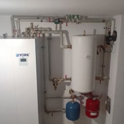 instal-bud Grzegorz Urbaniak - Biały piec CO marki York z podłączonymi rurami, zbiornikiem i osprzętem w kotłowni. Widoczne dwa zbiorniki wyrównawcze: niebieski i czerwony.