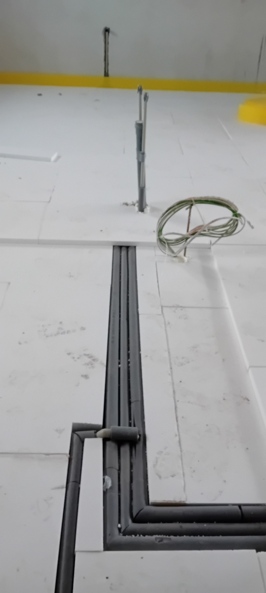 Białe płyty izolacyjne pod ogrzewanie podłogowe z widocznymi szarymi rurami instalacji. W tle pionowe rury i zwój kabli.