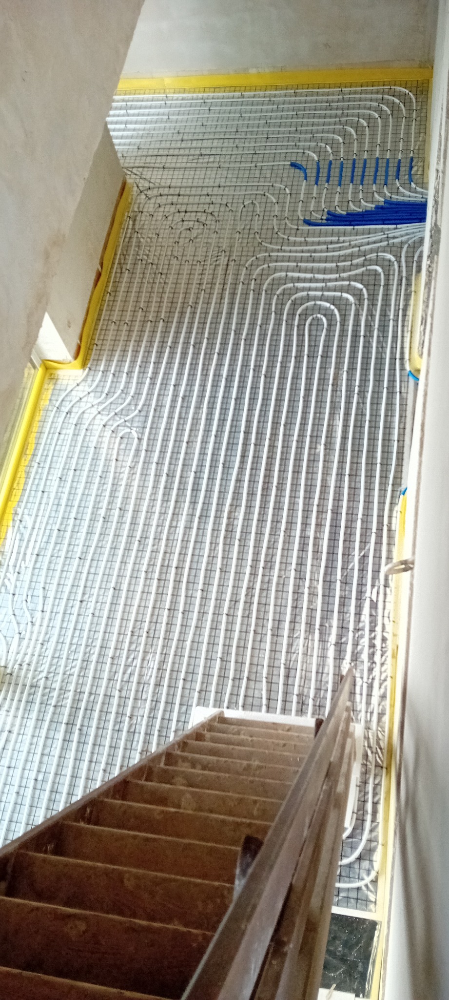 Białe rury ogrzewania podłogowego ułożone na siatce, widoczne z góry, z fragmentem drewnianych schodów w dolnej części kadru. Żółta taśma na obrzeżach.