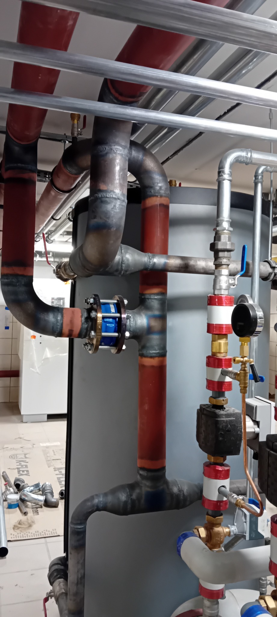 Złożona instalacja rur grzewczych z zaworami, manometrem i elementami łączeniowymi. Widoczne spawane łączenia i różnorodność materiałów. Część rur pomalowana na czerwono.