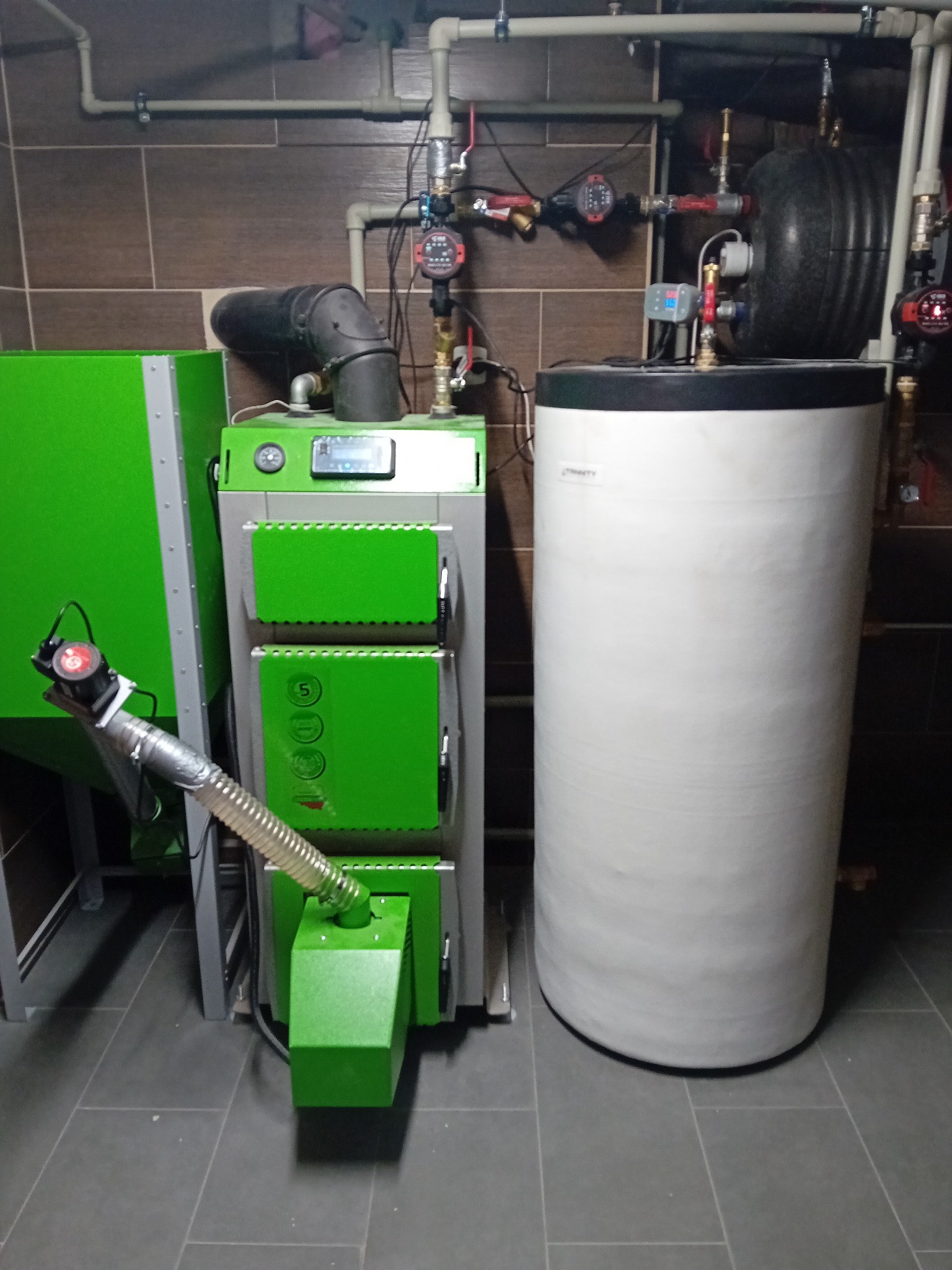 Nowoczesny piec CO na paliwo stałe, z podajnikiem i zasobnikiem, obok biały zbiornik akumulacyjny. Instalacja w kotłowni z brązowymi płytkami na ścianach.