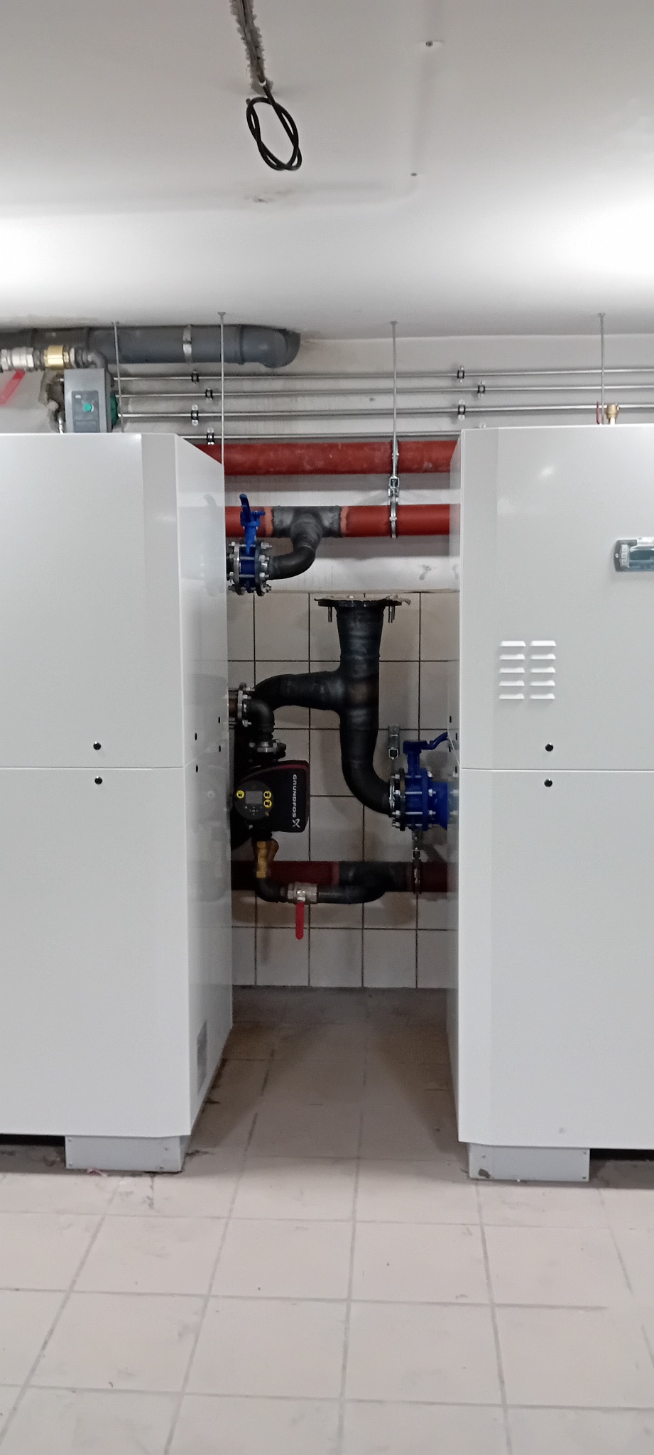 Nowoczesna kotłownia z dwoma białymi modułami i czarną instalacją rurową z niebieskimi zaworami. Widoczne rury w kolorze czerwonym i szarym pod sufitem.
