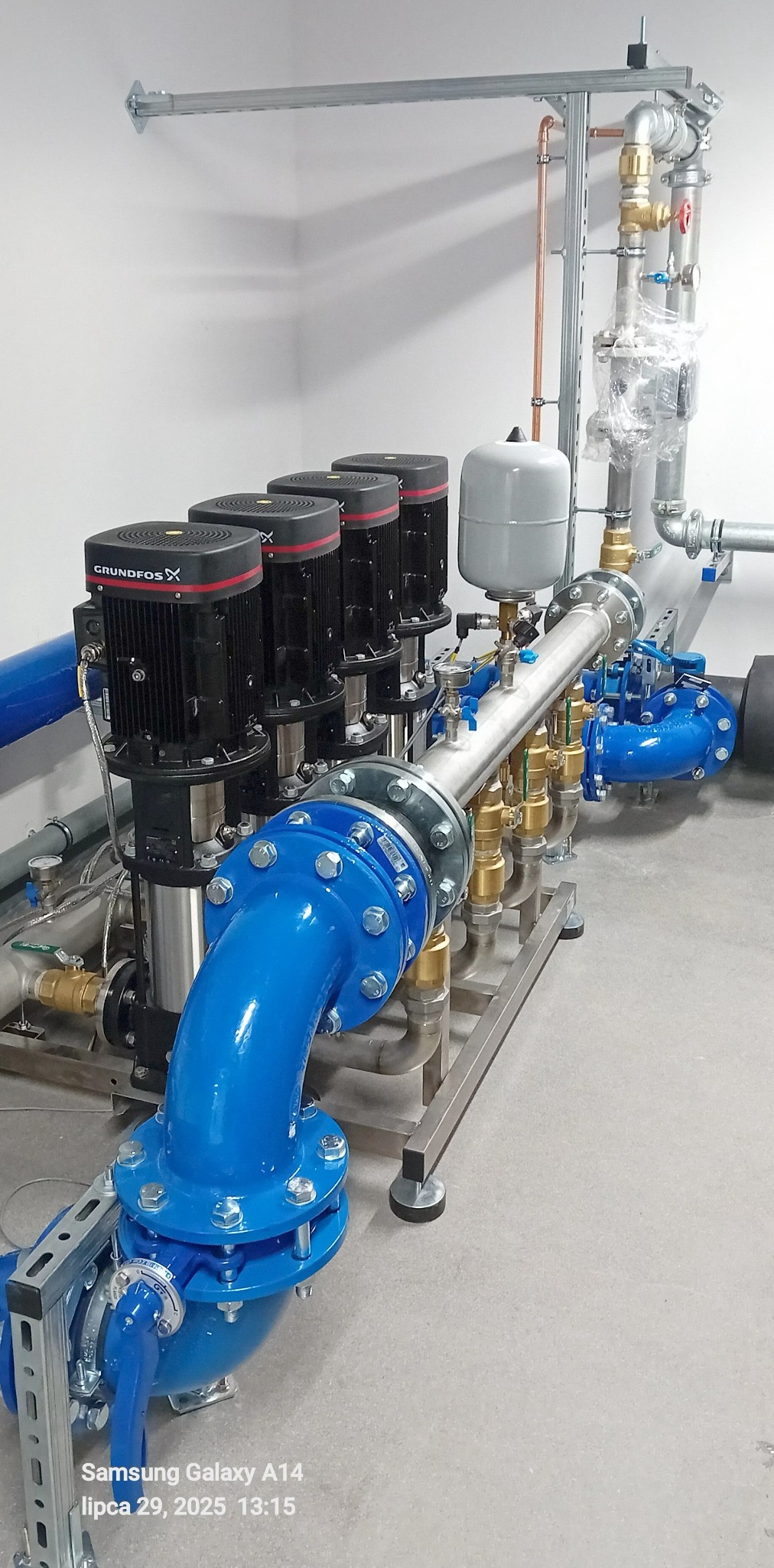 Złożona instalacja hydrauliczna z czterema pompami Grundfos, zbiornikiem wyrównawczym i rurami w kolorze niebieskim i miedzianym, zamontowana w jasnym pomieszczeniu.