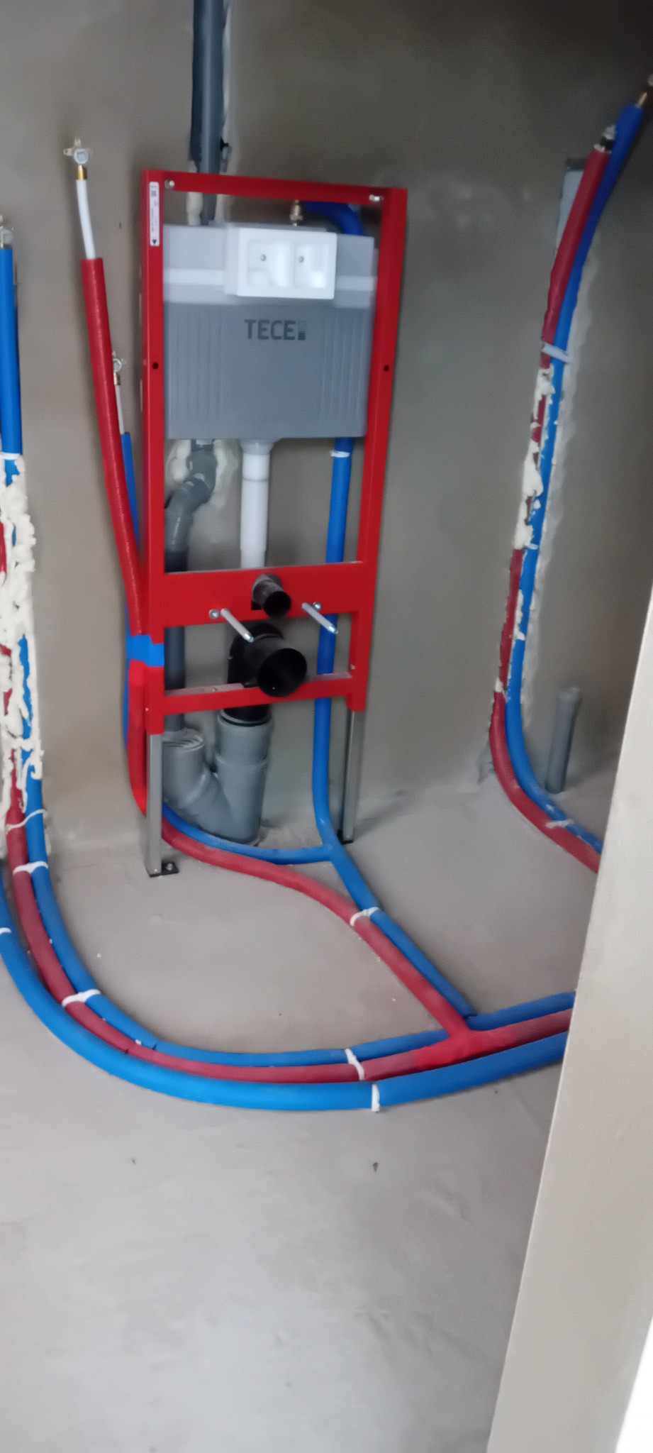 Stelaż podtynkowy TECE z instalacją wodną (rury PEX czerwone i niebieskie) w surowym pomieszczeniu, przed zabudową. Widoczne pianki montażowe.