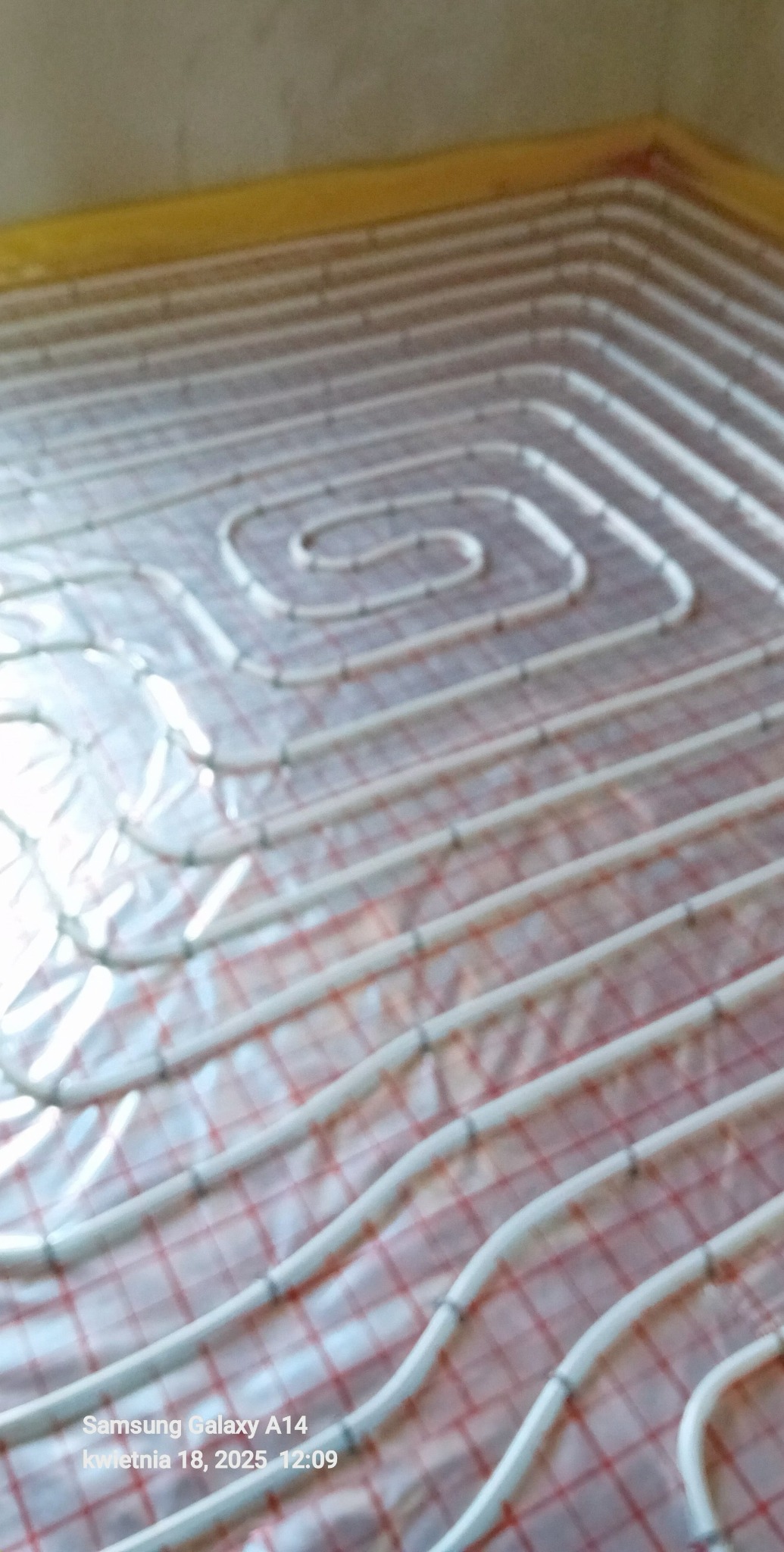 Białe rury ogrzewania podłogowego ułożone spiralnie na folii z czerwoną kratką, widoczne z bliska, z żółtą listwą przypodłogową w tle.