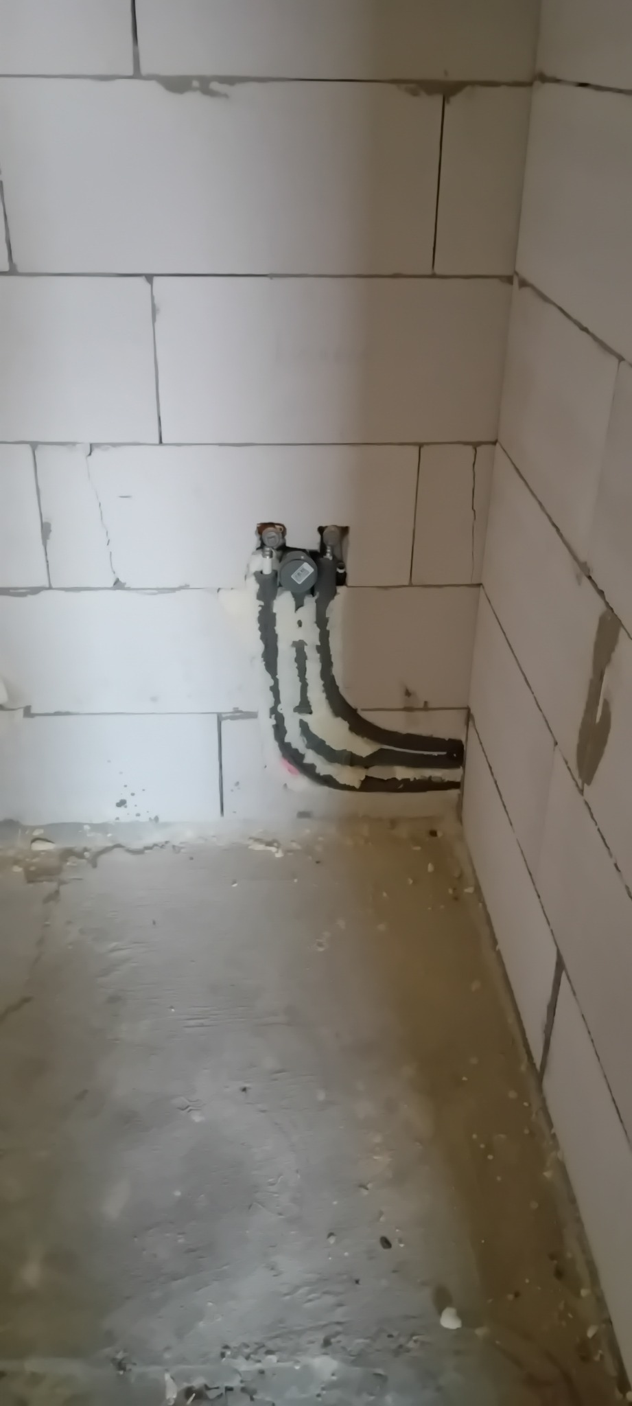 Fragment ściany z bloków, z widocznymi rurami instalacji hydraulicznej, częściowo zamurowanymi i uszczelnionymi, w surowym stanie wykończenia.